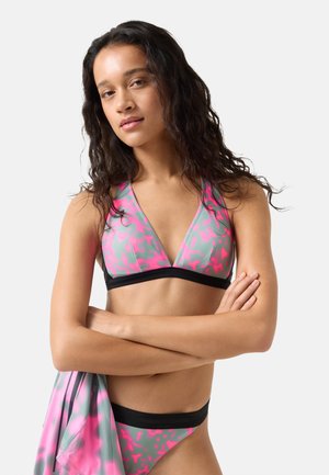 Femme aux longs cheveux bouclés portant un haut et un bas de bikini à motifs rose et gris, tenant un vêtement assorti, debout les bras croisés.