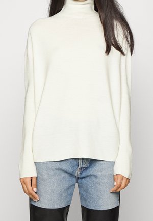 Femme portant un pull à col roulé blanc et un pantalon bicolore avec du denim clair au-dessus de panneaux en cuir noir.