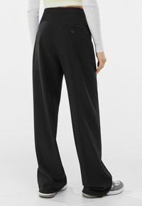Černé kalhoty širokého střihu s hladkým povrchem, vybavené malou zadní kapsou a detailním knoflíkem, v kombinaci s světle barevnými botami.