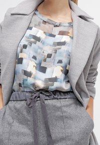 Blazer grigio su una blusa a fantasia con tonalità blu, nera e beige. Pantaloni grigi a vita alta con elastico in vita.