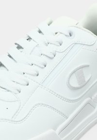 Weißer Ledersneaker mit flachen Schnürsenkeln, strukturierten Oberflächen, einem eingeprägten Logo und dezenten Steppnähten. Abgerundete Spitze und gepolsterte Sohle.