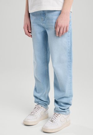 Persona che indossa jeans larghi azzurri, sneakers bianche e una camicia bianca in piedi davanti a uno sfondo semplice.