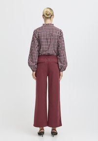 Bluză cu carouri, mâneci lungi, talie elastică și pantaloni largi bordeaux. Pantofi cu toc negri completează ținuta, văzută din spate.