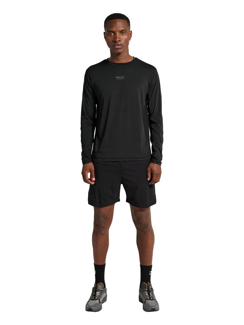 Homme debout portant un t-shirt à manches longues noir, un short noir, des chaussettes noires et des chaussures de sport grises sur fond blanc.