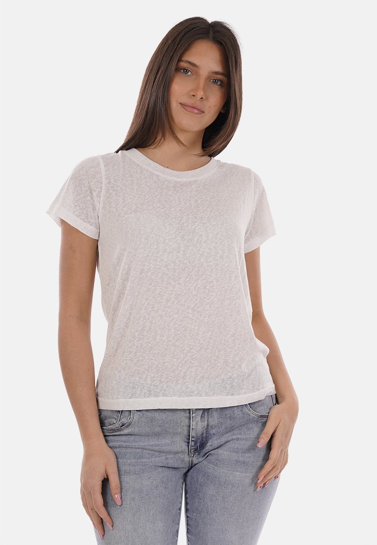 Jeune femme aux cheveux bruns et raides, portant un t-shirt blanc semi-transparent à manches courtes et un jean bleu clair, debout avec les mains détendues.