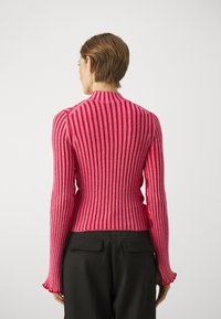 Sweter prążkowany w różnych odcieniach różu z długimi rękawami, wysokim kołnierzem i rozszerzanymi mankietami, zestawiony z czarnymi spodniami.