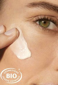 Produit de soin crémeux et blanc appliqué sur une peau claire. Texture lisse visible sur la joue, avec un œil vert en arrière-plan.