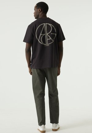 Man staat met zijn rug naar ons toe, draagt een zwart T-shirt met een groot rond ABG-logo, een donkere broek en witte sneakers op een egale achtergrond.