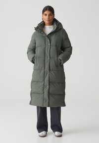 Lange, gevoerde jas in olijfgroen. Voorzien van een capuchon, drukknoopsluiting en een quilted textuur. Gekleed met donkere jeans en witte sneakers.