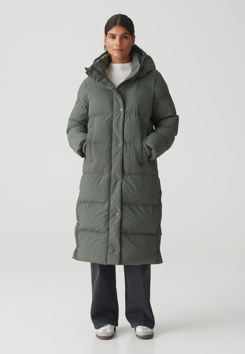 Lange, gevoerde jas in olijfgroen. Voorzien van een capuchon, drukknoopsluiting en een quilted textuur. Gekleed met donkere jeans en witte sneakers.