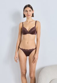 Rudos spalvos nėrinių ir audinio apatinių komplektas su švelniai paminkštinta liemenėle, dekoruota nėriniais, ir derančiomis kelnaitėmis-stringais, pasižyminčio minimalistišku dizainu.