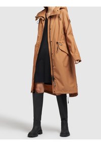 Parka marrone resistente all'acqua con cappuccio, chiusura frontale con zip, vita regolabile con coulisse e tasche laterali, abbinata a stivali neri fino al ginocchio.