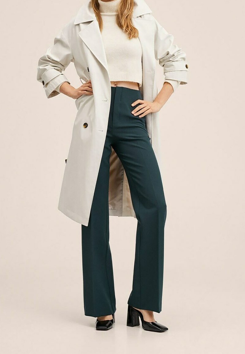 Femme portant un trench beige sur un pull col roulé court blanc, un pantalon large taille haute vert foncé et des talons blocs noirs.