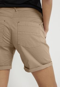 Pantalones cortos de algodón beige con puños enrollados, que cuentan con dos bolsillos traseros y sutiles detalles de costura. Textura suave con un ajuste relajado.