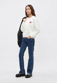 Sudadera blanca con un gráfico de labios rojos, combinada con jeans acampanados azul oscuro. La modelo lleva una chaqueta acolchada negra colgada sobre un hombro.