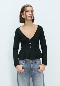 Crna pripijena cardigan jakna s dugim rukavima, V-izrezom i zatvaranjem na dugmad. Peplum dizajn u struku, upotpunjena svijetlim trapericama.