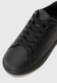 Svart lädersneaker med rund tå, strukturerad yta, platta svarta snörningar och logotyp på sidan med subtila perforeringar.