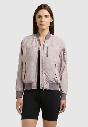 Kobieta stojąca przodem, ubrana w jasnoróżową kurtkę bomberkę na czarnych szortach na jednolitym tle.