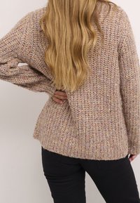 Pull en maille épaisse dans des tons pastel de rose et de beige avec un motif texturé. Il possède de longues manches et une coupe décontractée, arrivant au niveau des hanches.
