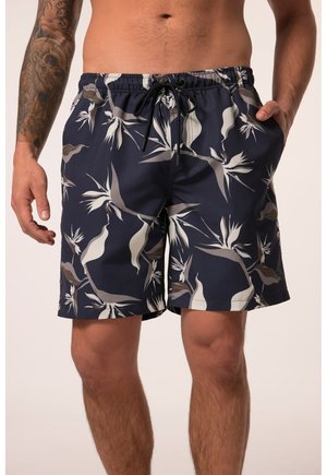 Mand iført marineblå badeshorts med hvide og grå tropiske bladmønstre og en sort snor, hånd i lommen, tatovering på venstre arm.