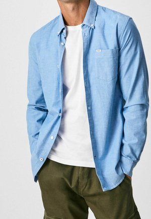 Mann trägt hellblaues Hemd über weißem T-Shirt und olivgrüne Hose, Hände in den Taschen vor einfarbigem Hintergrund.