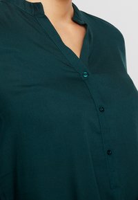 Teal skjorte med ståkrave iført af en person, der viser den øvre bryst- og nakkeområde mod en ensfarvet baggrund.