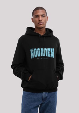 Young man wearing black hoodie with blue "NOORDEN" text and blue jeans, standing against plain light gray background.
