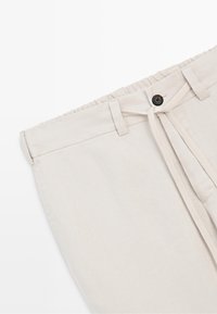 Beige trousers with an elastic waistband, button closure, belt loops, and a drawstring tied in a bow at the front.