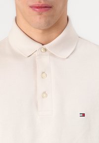 Polo beige avec un motif texturé, trois boutons et un petit logo brodé aux couleurs rouge, blanc et bleu sur la poitrine gauche.