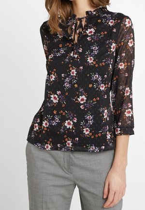 Sort gennemsigtig bluse med flerfarvet blomsterprint, lange ærmer og bindebånd ved halsen, båret med lysegrå ternede bukser.