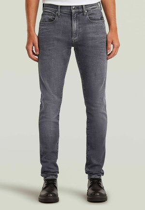 Mann trägt schlanke, ausgewaschene graue Jeans kombiniert mit schwarzen Leder-Schnürstiefeln, steht vor einem einfarbigen hellen Hintergrund.