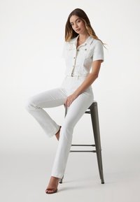 Bílá denní bunda s krátkými rukávy a zapínáním na knoflíky, kombinovaná s bílými džínami s vysokým pasem a zvonovým střihem. Modelka sedí na šedé barové židli.