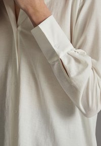 Ljusbeige knapp-up skjorta med långa ärmar, med detaljer vid manschetterna och ett mjukt, texturerat tyg. Knapparna är synliga på framsidan.