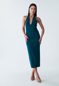 Teal halter-nekkimekkoinen mekko, jossa on syvä V-pääntie, tiukka siluetti ja polvipituus. Hengittävä kangas, jossa on hieno tekstuuri; yhdistettynä nude-sandaaleihin.