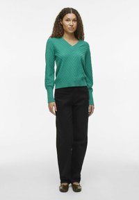 Grüner V-Ausschnitt-Pullover mit einem Zickzack-Muster, kombiniert mit schwarzen, locker sitzenden Hosen und dunklen flachen Schuhen. Das Model steht vor einem neutralen Hintergrund.