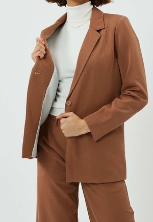 Osoba ubrana w brązowy garnitur z marynarką na jeden guzik, pod którą ma biały golf, trzymająca marynarkę otwartą jedną ręką.