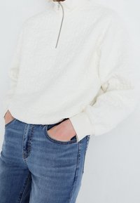 Sudadera blanca acolchada con media cremallera y cuello alto, combinada con unos vaqueros de mezclilla azul claro; presenta una textura suave y un corte relajado.