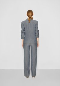 Donna con i capelli raccolti, che indossa un blazer grigio a righe sottili ben aderente e pantaloni a gamba larga abbinati, in piedi contro uno sfondo chiaro e uniforme.