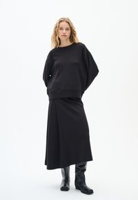 Felpa nera oversize e gonna lunga realizzata in un tessuto liscio. Abbinata a stivali neri fino al ginocchio. Design e silhouette minimalisti.