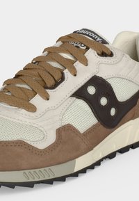 Κοντινή λήψη ενός μπεζ και καφέ sneaker Saucony με υφασμάτινα καφέ κορδόνια και ορατό ανώτερο μέρος από δίχτυ.