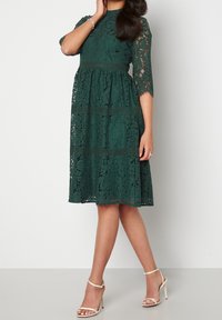 Robe en dentelle verte avec manches trois-quarts, corsage ajusté et jupe évasée. Présente des motifs floraux et un ourlet longueur thé.