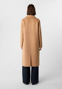 Persona in piedi con la schiena rivolta, indossa un lungo cappotto color cammello sopra pantaloni larghi scuri, contro uno sfondo chiaro e neutro.