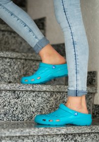 Türkise Holzschuhe mit Belüftungsöffnungen und strukturiertem Obermaterial. Getragen mit hellblauen, eng anliegenden Jeans beim Betreten grauer Steinstufen.