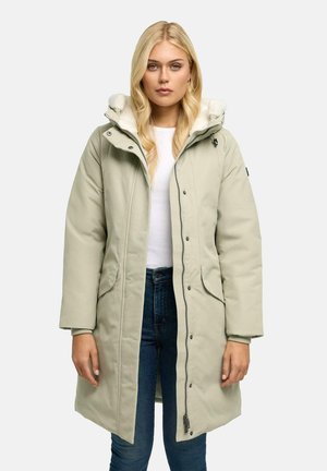 Kvinde iført en lang lysegrøn vatteret frakke med en hvid fleeceforet hætte, hvid skjorte og blå jeans, stående mod en ensfarvet baggrund.
