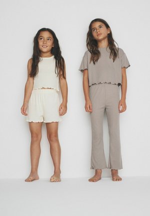 Dve bosé dievčatá stoja, jedna má krémový tielko bez rukávov a krátke nohavice, druhá hnedošedý krátky rukáv a nohavice, oproti bielemu múru.