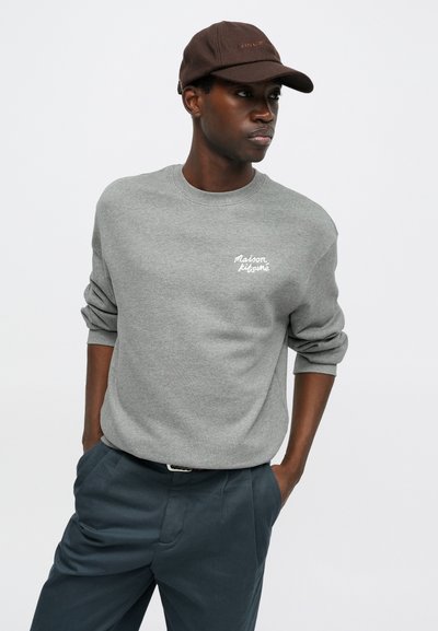 Halli sweatshirt ümmarguse kaelusega ja volditud varrukatega, millel on valge tikandtekst; koos tumesiniste pükste ja pruuni mütsiga.