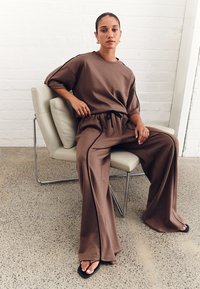 Top beige oversize e pantaloni a gamba larga in tessuto morbido, con vita regolabile e strisce laterali a contrasto. Indossati con ciabatte nere.