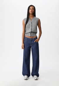 Šedý bezrukávový košili na knoflíky vyrobený z pletené tkaniny, kombinovaný s tmavými džínami s vysokým pasem a širokým střihem; model má na sobě stříbrné tenisky.