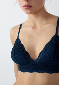 Bralette en dentelle marine de forme triangulaire, avec des bords festonnés, des motifs floraux et des fines bretelles. La texture douce améliore le design.