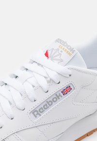 Białe skórzane sneakersy z płaskimi sznurówkami, wyściełanym kołnierzem, logo Reebok i detalem w stylu Union Jack. Posiadają teksturowaną gumową podeszwę.